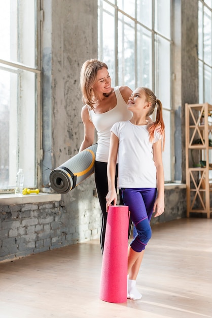 Stuoie di yoga della tenuta della figlia e della foto a figura intera che se lo esaminano