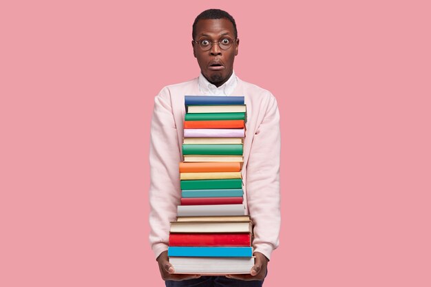 Stupefatto giovane afroamericano con una grande pila di libri, vestito con un maglione casual, ha sorpreso l'espressione del viso