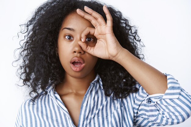 Stupita affascinante giovane ragazza millenaria dalla pelle scura con i capelli ricci bocca aperta suono wow che mostra gesto ok ok guardando attraverso cirlce stupito, impressionato, studio muro bianco