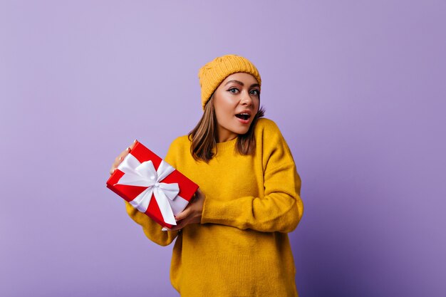 Stupita ragazza attraente in abito giallo in posa con regalo di compleanno. Donna di buon umore in maglione elegante divertendosi a Natale.