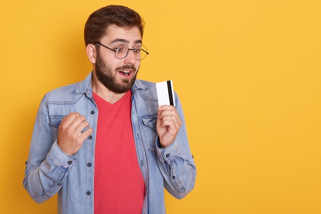 Stupito uomo con la barba che indossa giacca di jeans, camicia rossa e occhiali, con in mano la carta di credito e sembra una carta con la bocca aperta