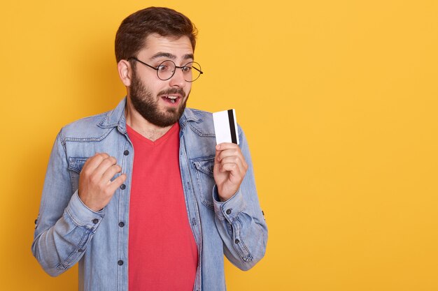 Stupito uomo con la barba che indossa giacca di jeans, camicia rossa e occhiali, con in mano la carta di credito e sembra una carta con la bocca aperta