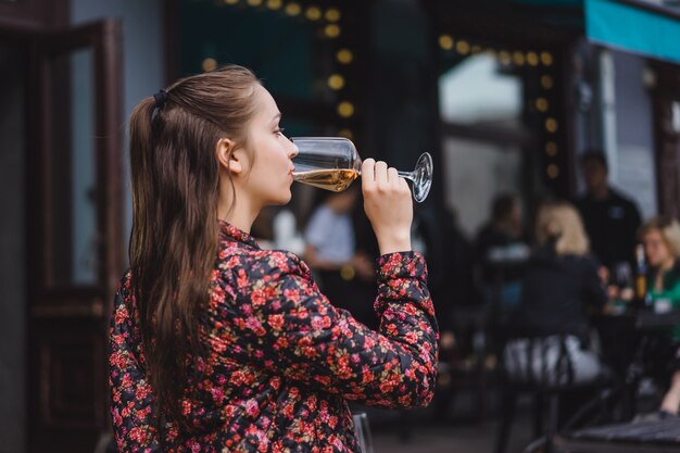 Stylish young girl beve vino in una caffetteria in una terrazza estiva. Una ragazza con i capelli lunghi gode di un bicchiere di vino in una serata estiva. Ritratto. avvicinamento