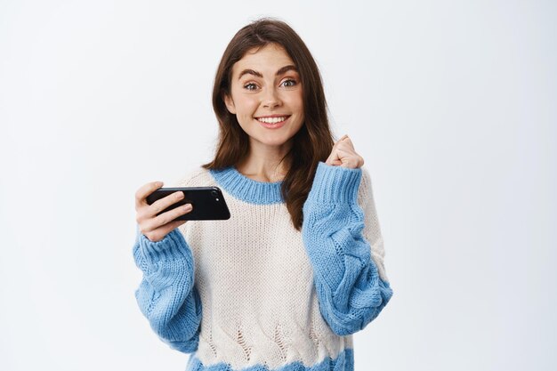 Successo. Eccitato giovane donna che gioca al videogioco mobile, tenendo lo smartphone in orizzontale e sorridendo divertito, vincendo online, in piedi in un maglione su bianco