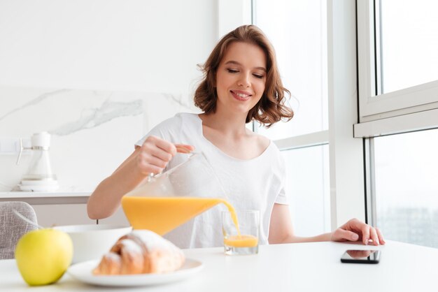 Succo di versamento della donna castana allegra in vetro mentre sedendosi e facendo colazione alla cucina