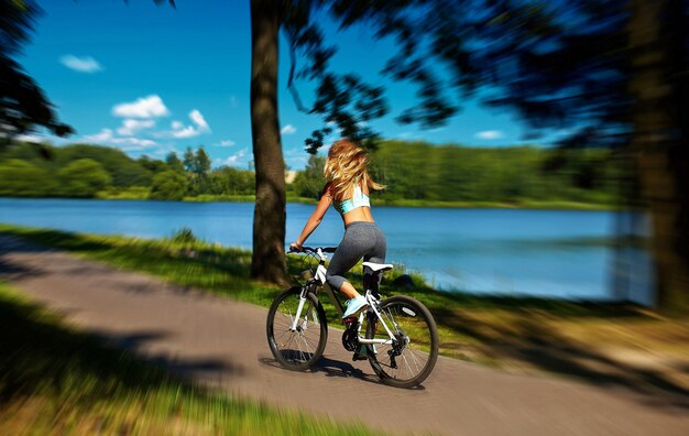 Sul retro del modello sexy della ragazza della donna bionda di sport caldo che guida sulla bicicletta nel parco verde di estate vicino al lago con il volo ha elevato i capelli in aria