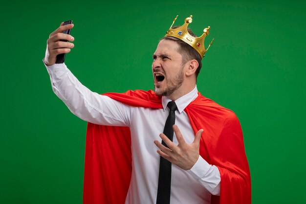 Super eroe imprenditore in mantello rosso che indossa la corona facendo selfie utilizzando lo smartphone impazzendo in piedi arrabbiato selvaggio su sfondo verde