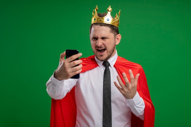 Super eroe uomo d'affari in mantello rosso che indossa la corona utilizzando smartphone guardando lo schermo impazzendo in piedi arrabbiato selvaggio su sfondo verde