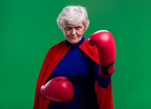 Supereroe donna anziana che indossa mantello rosso con guantoni da boxe che guarda l'obbiettivo con espressione seria e sicura pronta a combattere