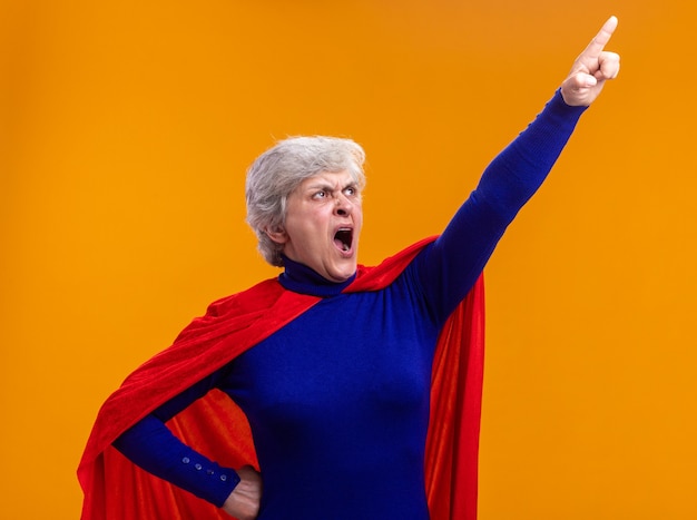 Supereroe donna anziana che indossa un mantello rosso che guarda in alto puntando con il dito indice a qualcosa che grida con un'espressione aggressiva in piedi su sfondo arancione
