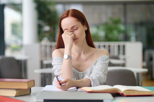 Superlavoro. Giovane donna stanca con lunghi capelli rossi con gli occhi chiusi che si toccano mano a faccia seduta a tavola con libri in una stanza luminosa