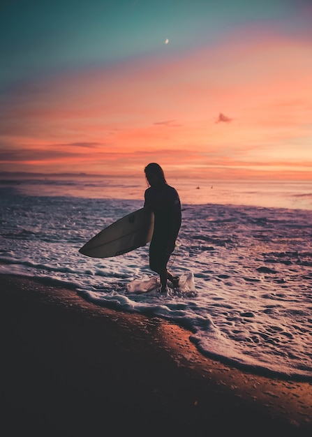 Surfista con una tavola che esce dal mare durante il tramonto