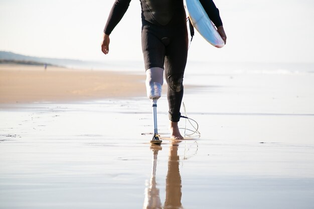 Surfista irriconoscibile con protesi della gamba che cammina vicino al mare