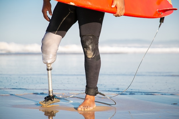 Surfista maschio ritagliata in piedi con la tavola da surf sulla spiaggia del mare