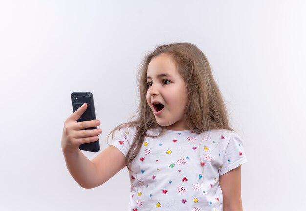 T-shirt bianca da portare sorpresa della bambina della scuola che osserva sul telefono su fondo bianco isolato