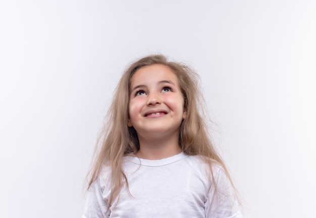 T-shirt bianca da portare sorridente della bambina della scuola che osserva in su su fondo bianco isolato