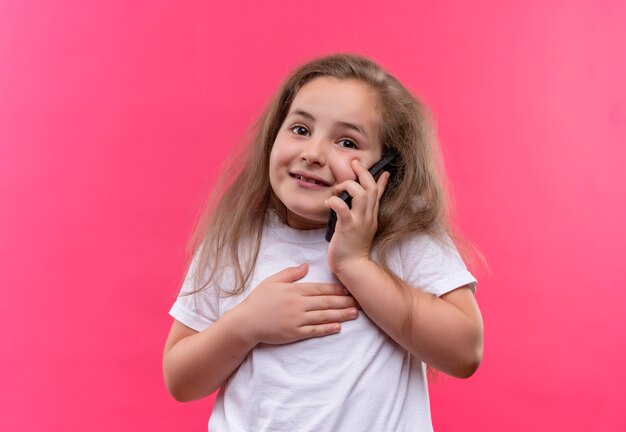 T-shirt bianca da portare sorridente della bambina della scuola parla sul telefono su fondo rosa isolato