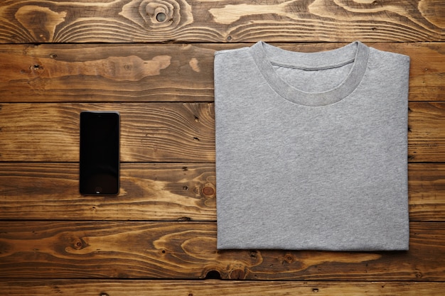 T-shirt grigia in bianco accuratamente piegata vicino al gadget smartphone nero sulla vista dall'alto del tavolo in legno rustico