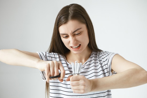 Tabacco, fumo e concetto di stile di vita sano. Forte bella giovane femmina caucasica avvilita in top a strisce che taglia molte sigarette usando le forbici, sentendosi pronta a rompere finalmente la cattiva abitudine