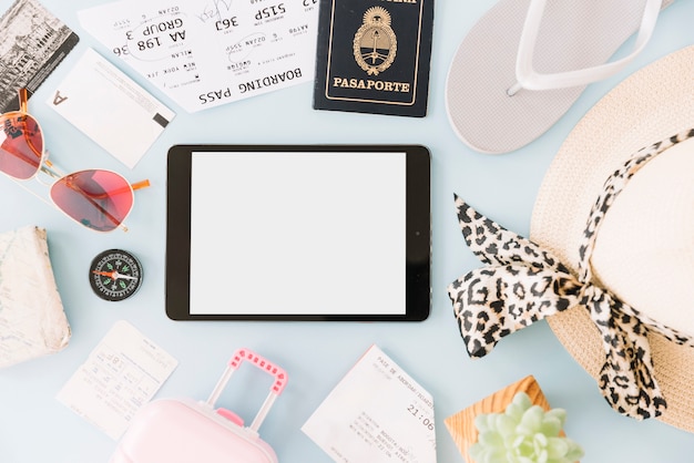 Tablet digitale vuoto circondato da carte d&#39;imbarco; biglietto da visita; occhiali da sole; bussola; pianta di cactus; cappello; passaporto; borsa da viaggio in miniatura e infradito