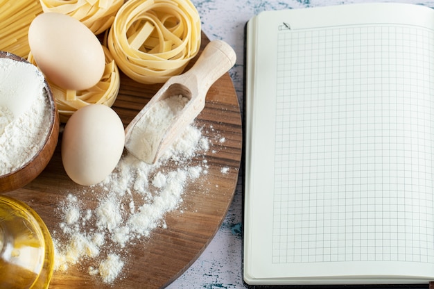 Tagliatelle con olio, uovo e ciotola di farina su tavola di legno e quaderno.