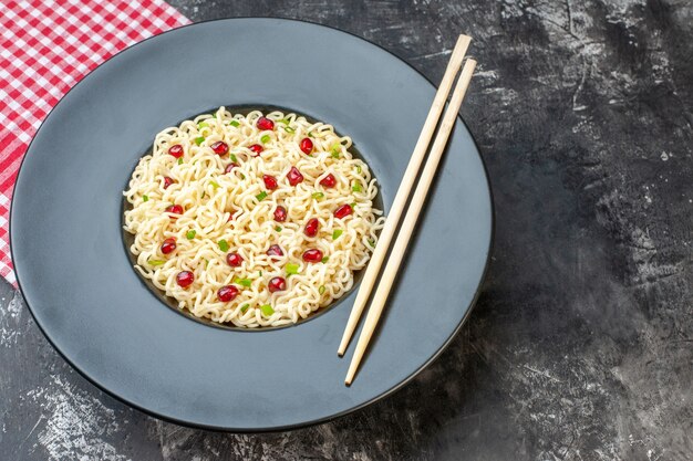 Tagliatelle di ramen di vista dal basso con le bacchette dei melograni sul tovagliolo a quadretti bianco rosso del piatto rotondo scuro sulla tavola scura