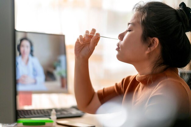 Tampone nasale asiatico della mano femminile che testa i test rapidi da sola per il rilevamento del virus SARS co2 mediante istruzioni del medico televideo a casa isolare il concetto di quarantena