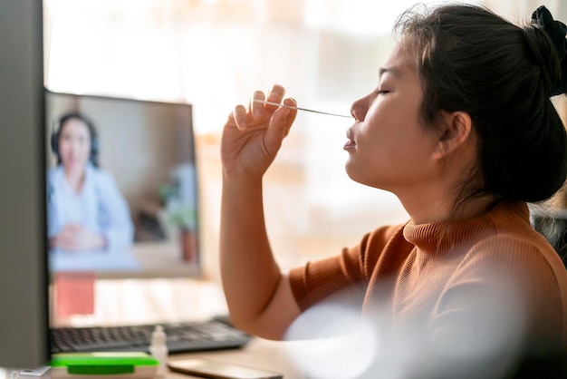 Tampone nasale asiatico della mano femminile che testa i test rapidi da sola per il rilevamento del virus SARS co2 mediante istruzioni del medico televideo a casa isolare il concetto di quarantena