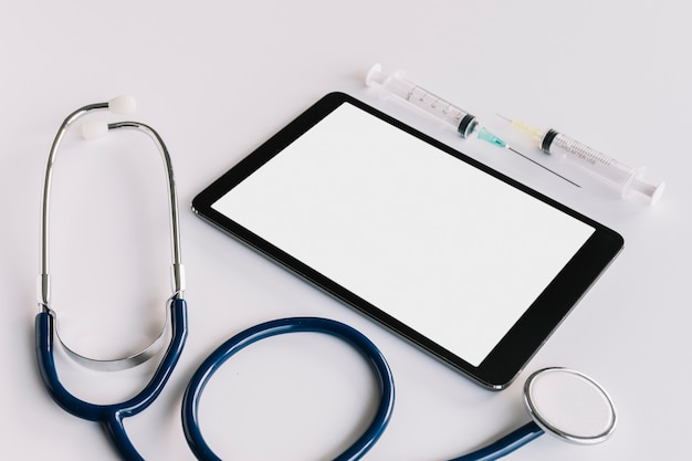 Tavoletta digitale con schermo vuoto; siringa e stetoscopio su sfondo bianco