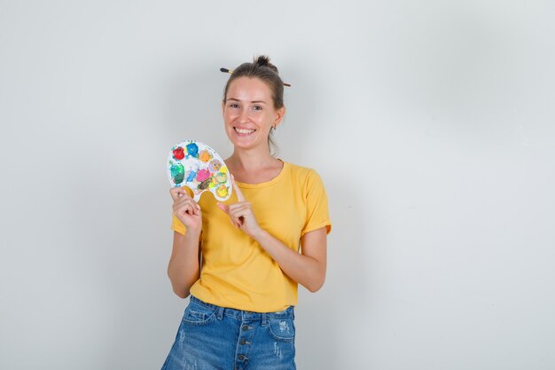 Tavolozza di arte della holding della giovane donna in maglietta gialla, pantaloncini di jeans e allegro