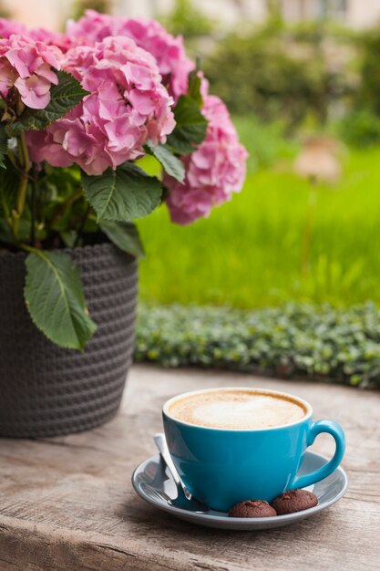 Tazza blu del caffè del latte con i biscotti sul vaso di fiore rosa vicino di superficie di legno