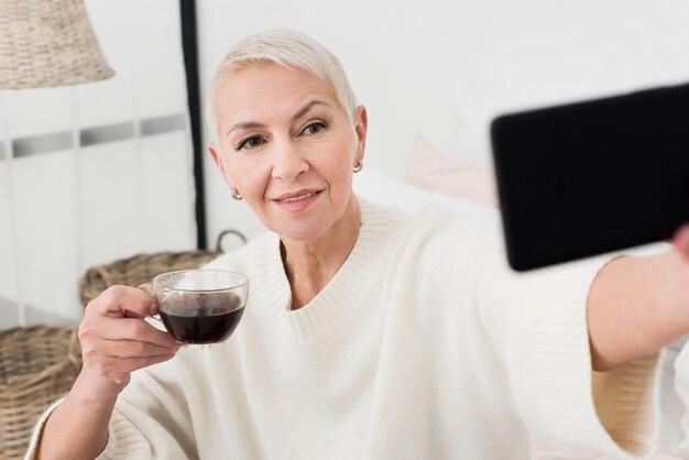 Tazza di caffè anziana della tenuta della donna e prendere selfie