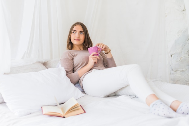 Tazza di caffè della tenuta della donna che si trova sul letto con il libro