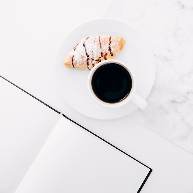 Tazza di caffè e croissant sul piatto vicino al taccuino pagina vuota