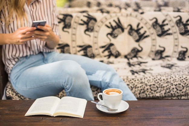 Tazza di caffè e libro aperto davanti alla donna che per mezzo del telefono cellulare