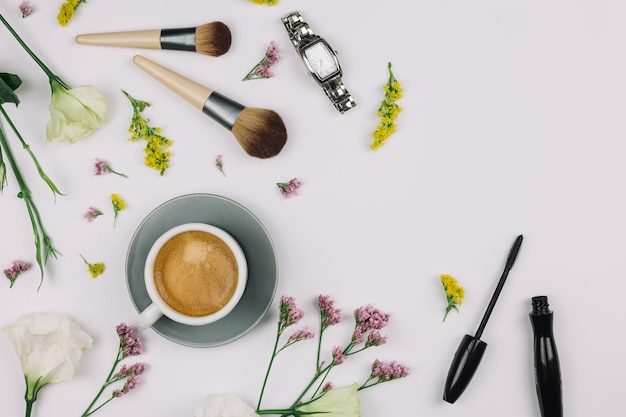 Tazza di caffè; orologio da polso; pennello da trucco; bottiglia di mascara con fiori freschi su sfondo bianco
