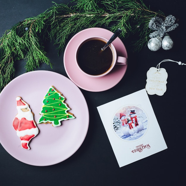 Tazza di caffè rosa, piatto con pan di zenzero e cartolina di Natale si trovano sul tavolo nero