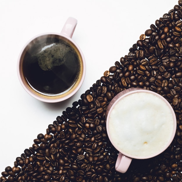 Tazza di caffè si leva in piedi sul tavolo bianco e tazza di latte sul tavolo coperto di grani di caffè