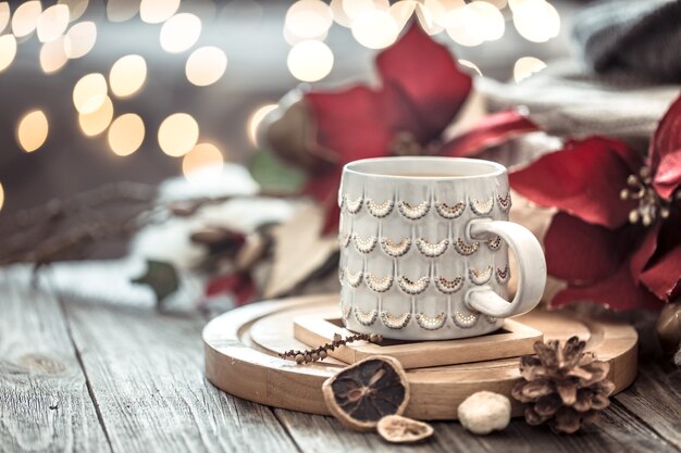 Tazza di caffè sopra il bokeh delle luci di Natale in casa sulla tavola di legno con i fiori su una parete e sulle decorazioni. Decorazione natalizia, magico Natale