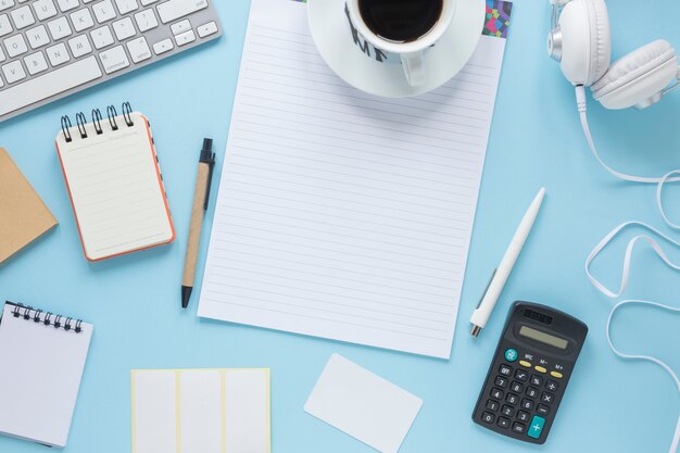 Tazza di caffè su una sola riga; blocco note a spirale; penna; tastiera; cuffia su sfondo blu