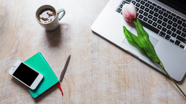 Tazza di caffè; tulipano sul laptop; cellulare; penna e libro sul tavolo di legno