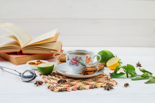 Tazza di tè con cannella e limone sulla tovaglietta quadrata con lime, una ciotola di mandorle, un colino da tè e libri sulla superficie bianca