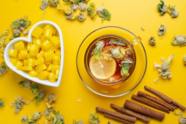 Tè al limone con erbe secche, zollette di zucchero, bastoncini di cannella in una tazza su superficie gialla, piatto disteso.