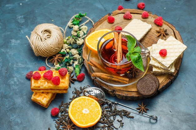 Tè con erbe, frutta, spezie, biscotti, colino, filo in una tazza su tavola di legno e sfondo di stucco, piatto.