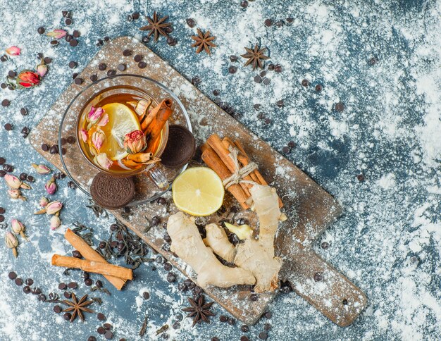 Tè in una tazza con farina, cioccolato, biscotti, spezie, limone, vista dall'alto di erbe su cemento e tagliere