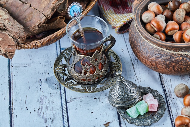Tè nero in una tazza di vetro tradizionale con caramelle e una ciotola di nocciole sul tavolo di legno blu