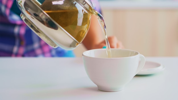 Tè verde che scorre dalla teiera al rallentatore. Primo piano di tè dal bollitore versare lentamente nella tazza di porcellana in cucina al mattino a colazione, utilizzando tazza da tè e foglie di erbe salutari.