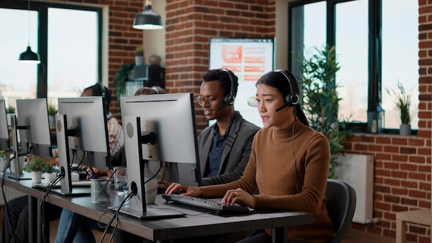 Team multietnico di persone che lavorano presso l'ufficio del call center, utilizzando cuffie audio per le telecomunicazioni per aiutare i clienti. Persone che rispondono alla chiamata sulla linea di assistenza telefonica, fornendo assistenza alla postazione di lavoro.