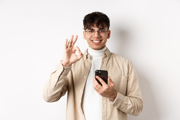 Tecnologia e concetto di acquisto online. Ritratto di un bel ragazzo moderno con gli occhiali che mostra il gesto OK usando lo smartphone, consigliando app o negozio, sfondo bianco