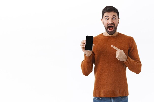 Tecnologia, persone e concetto di comunicazione. Ritratto di un uomo caucasico entusiasta e sorridente che punta il dito sullo schermo del telefono cellulare, guadagna bonus nell'app per smartphone online, sembra impressionato